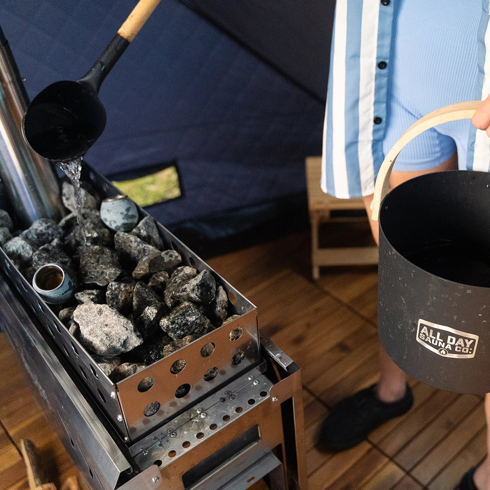 Up close view of sauna stove and rocks. Someone is pouring water over the sauna rocks inside the All Day Sauna Co. outdoor sauna tent